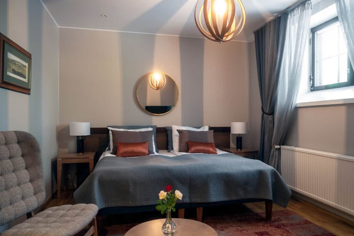 Een hotelkamer met een groot bed, twee nachtkastjes met lampen, een ronde spiegel, een stoel, een klein rond tafeltje met een bloevas en een raam met gordijnen.