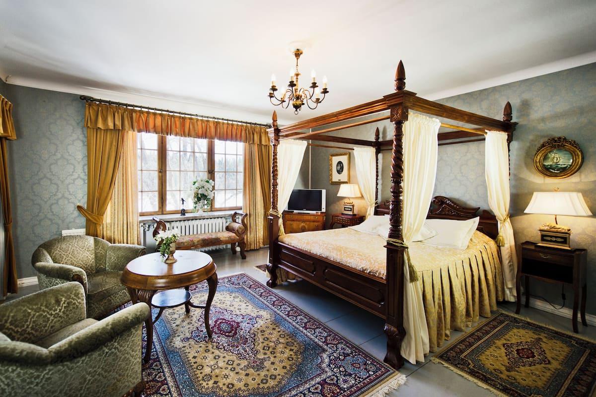 Een hemelbed met houten poten, een ronde houten tafel met een bloemenvaas, een bank en een raam met gordijnen.