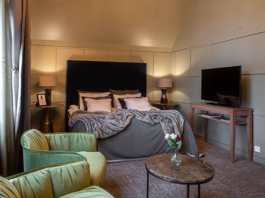 A hotel room with a bed, a television, a small table with a vase, and a green armchair.
