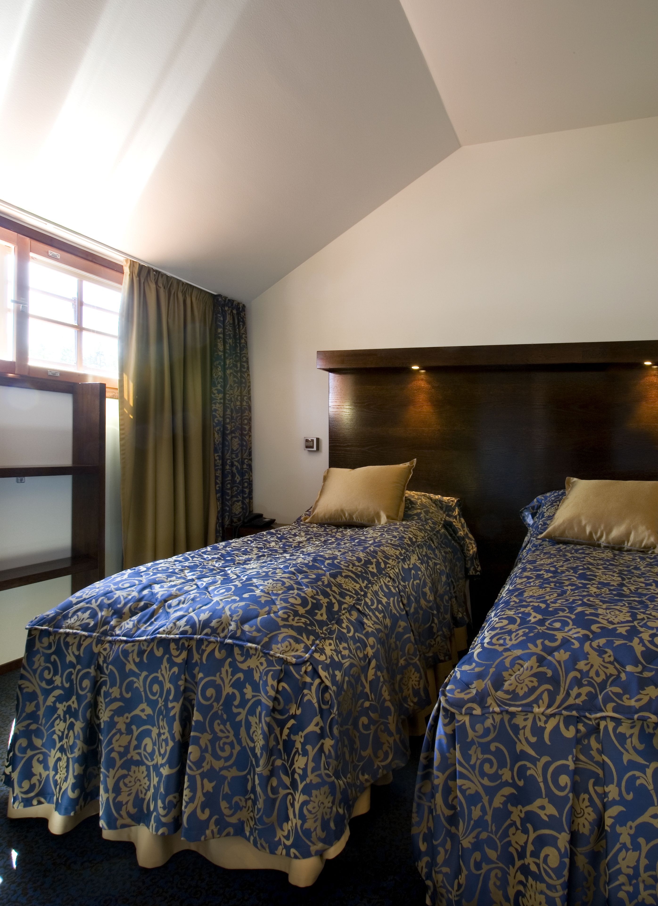 Ein Hotelzimmer mit zwei Betten, einem Holzkopfteil, einem Fenster mit Vorhängen und einem kleinen Nachttisch.
