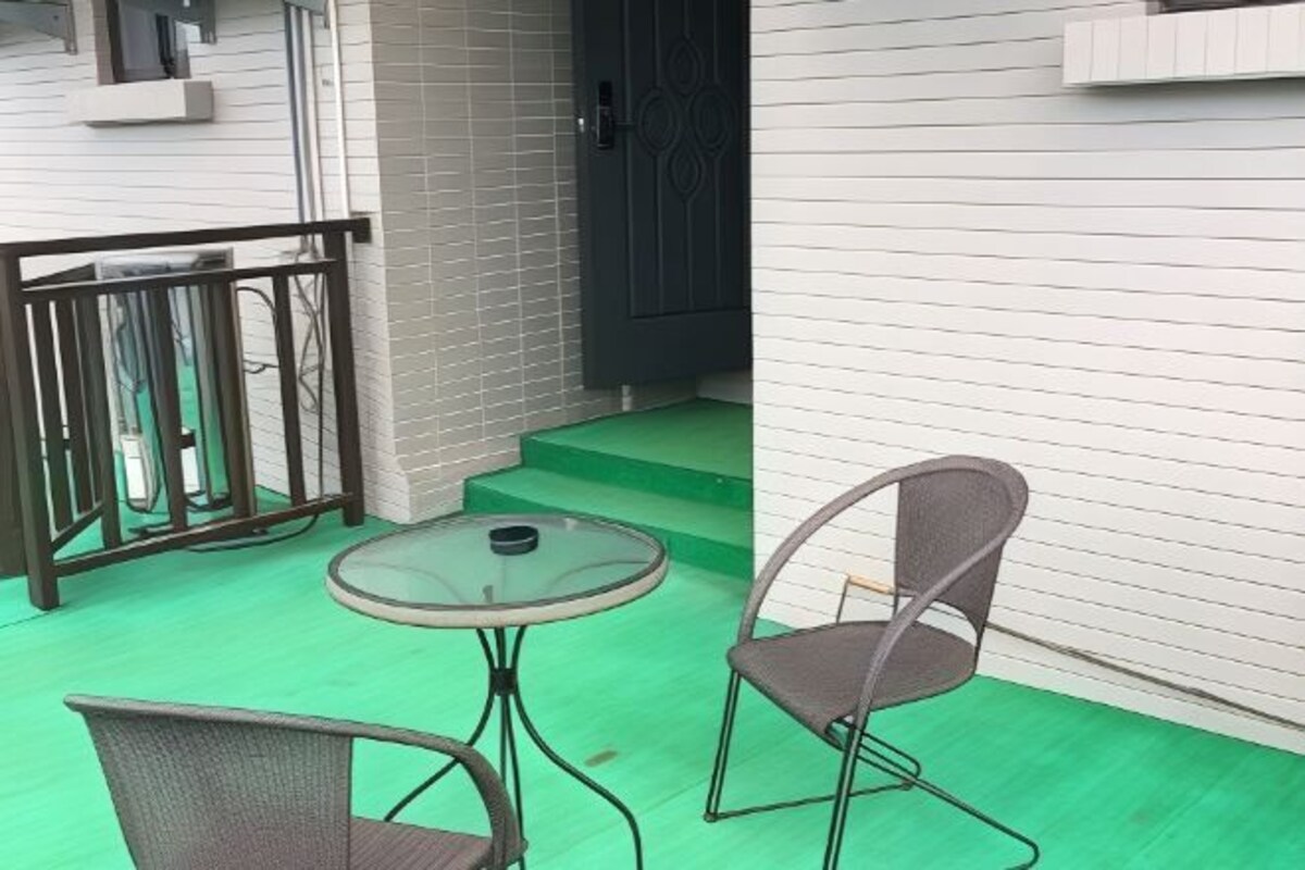 Una pequeña zona de estar al aire libre con una mesa redonda y dos sillas, una barandilla metálica y piso verde.