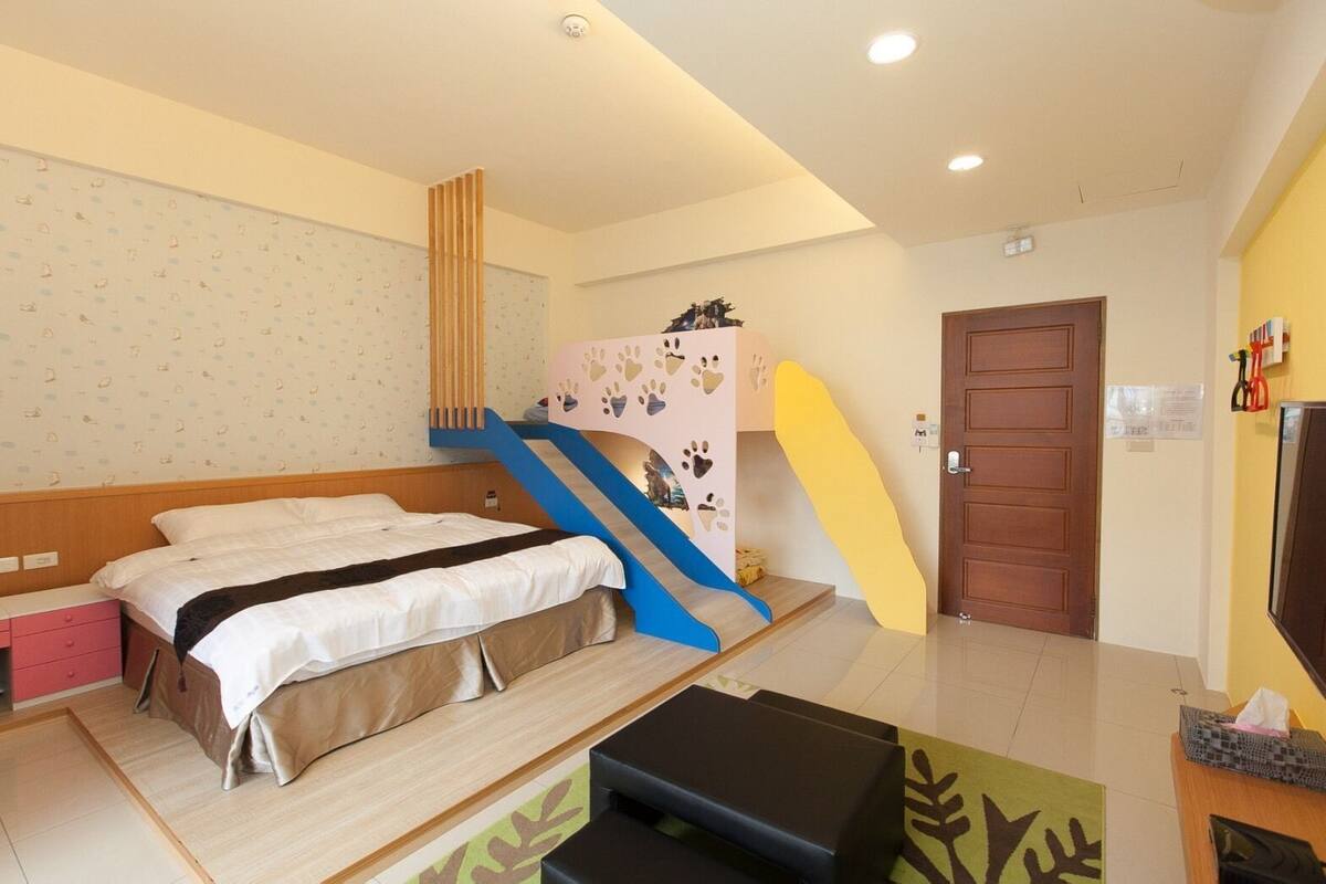A hotel room with a bed, a desk, a chair, a TV, a door, and a staircase with a colorful design.