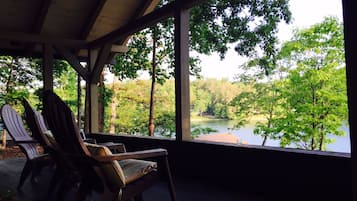 Un espace extérieur aménagé avec une zone de repos couverte, des fauteuils Adirondack, une vue sur un lac entouré d’arbres et une terrasse en bois.