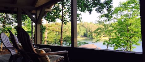 Un espacio de estar al aire libre con techo, sillas Adirondack, vista a un lago rodeado de árboles y una terraza de madera.