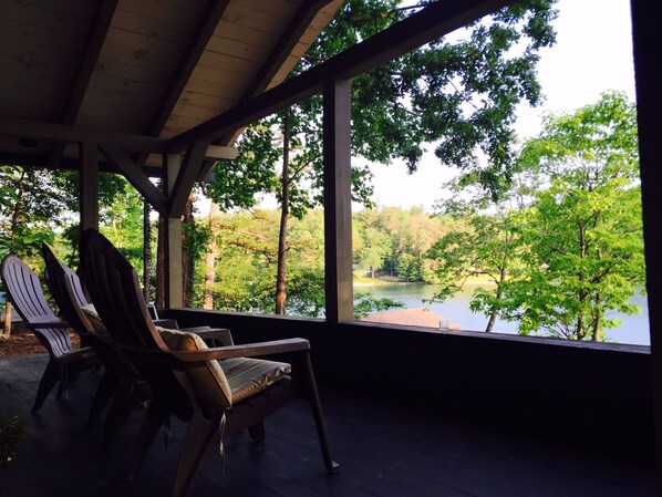 A covered outdoor seating area with Adirondack chairs, a view of a lake surrounded by trees, and a wooden deck.