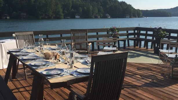 A wooden deck with a table set for dining, overlooking a lake with a forested shoreline.