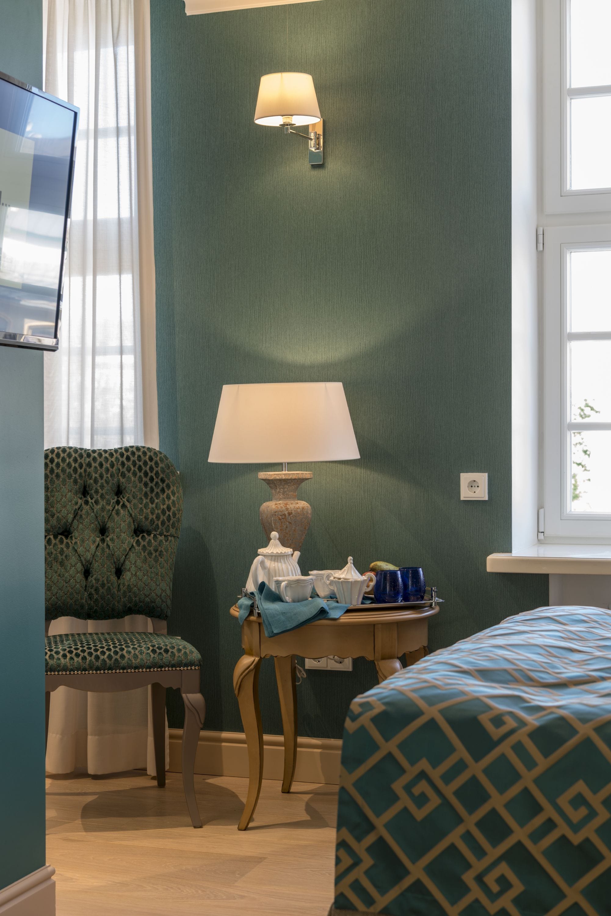 A hotel room with a green wall, a tufted armchair, a wooden nightstand with a lamp, a bed with patterned bedding, and a television on the left.