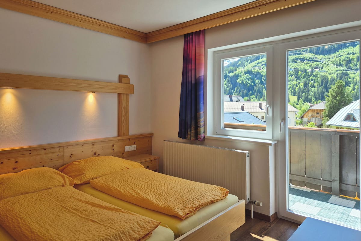 Une chambre à coucher avec une tête de lit en bois, un lit avec une literie jaune, une fenêtre donnant sur une zone boisée et un radiateur.