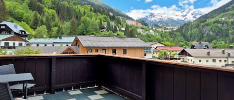 A balcony with a checkered floor, overlooking a mountainous landscape with snow-capped peaks and a village below.