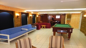 Taula de bitlles, dues cadires amb ratlles, un sofà blau i un canapè vermell en una habitació amb parets de fusta i paviment de rajola.