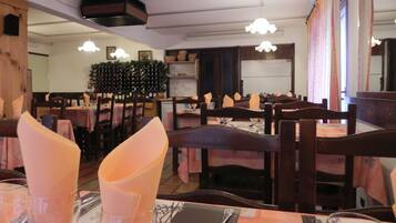 Un interno di ristorante con tavoli e sedie di legno, tavoli apparecchiati con cura e tovaglioli, e una cantina a vista sullo sfondo.