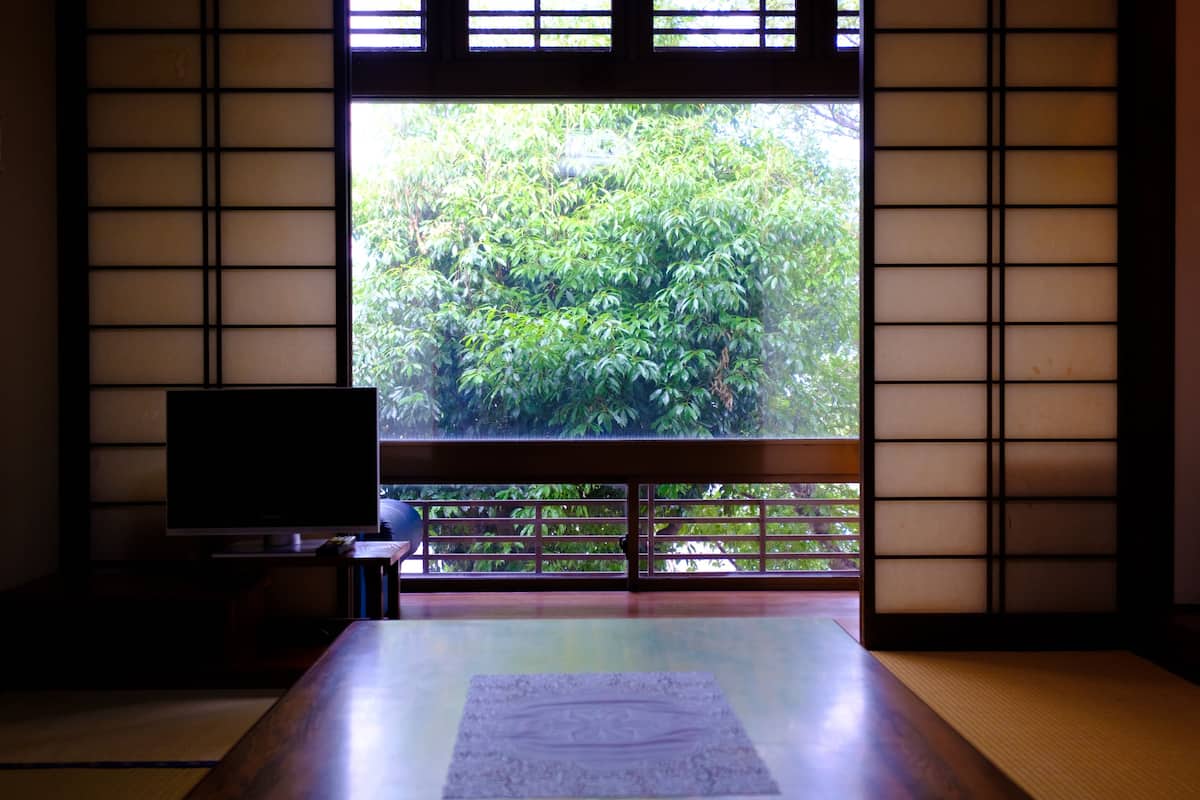A traditional Japanese room with tatami flooring, a television on a low table, and a view of lush greenery through shoji screens.