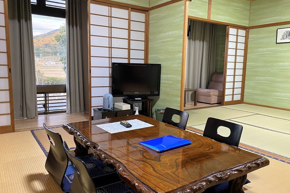 A traditional Japanese-style room with tatami flooring, a wooden table with chairs, a television, and a sofa.