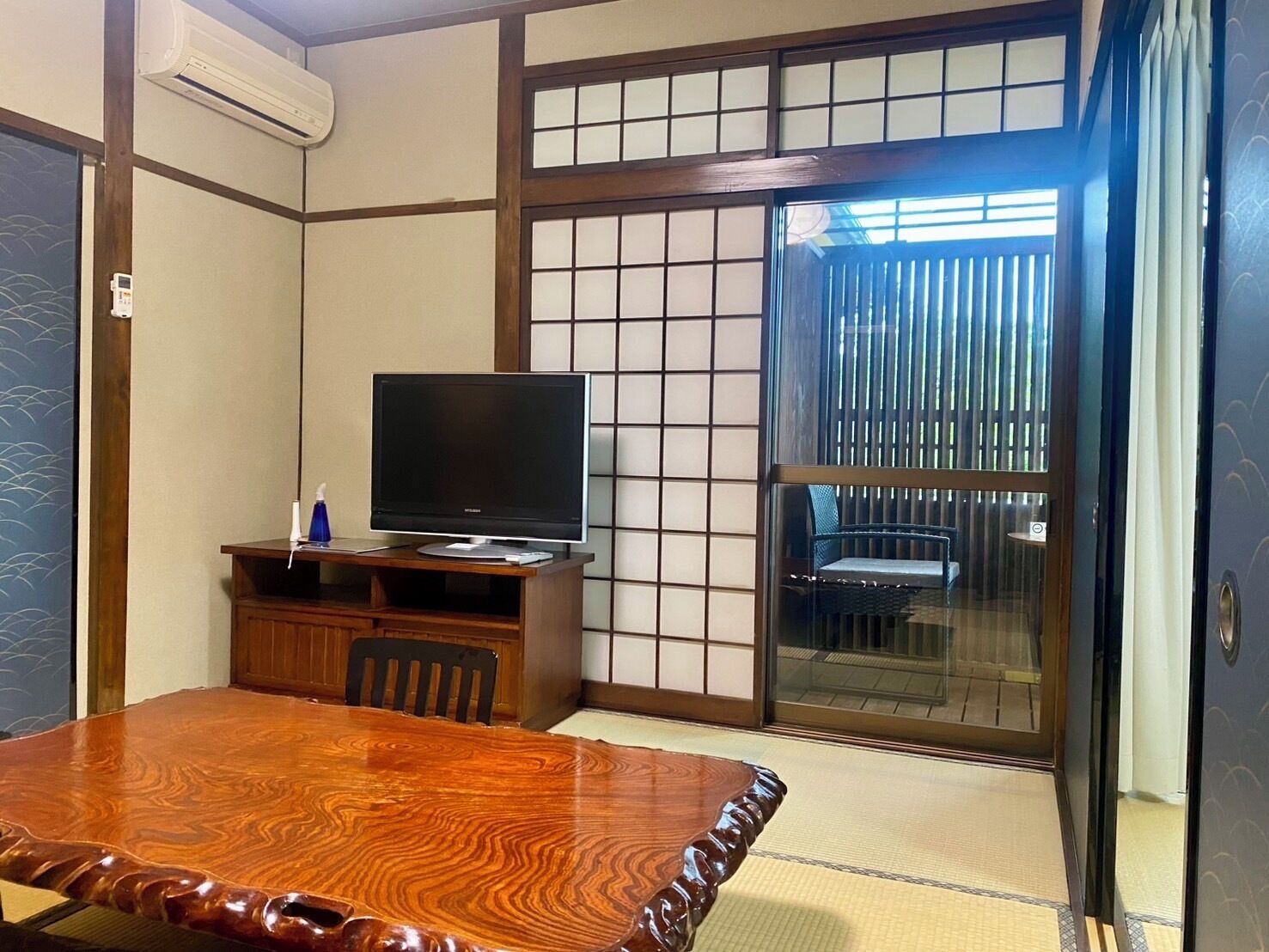 Une pièce de style japonais traditionnel, avec un sol recouvert de tatamis, une table en bois, un téléviseur posé sur un meuble bas et une porte-fenêtre coulissante donnant sur un balcon.