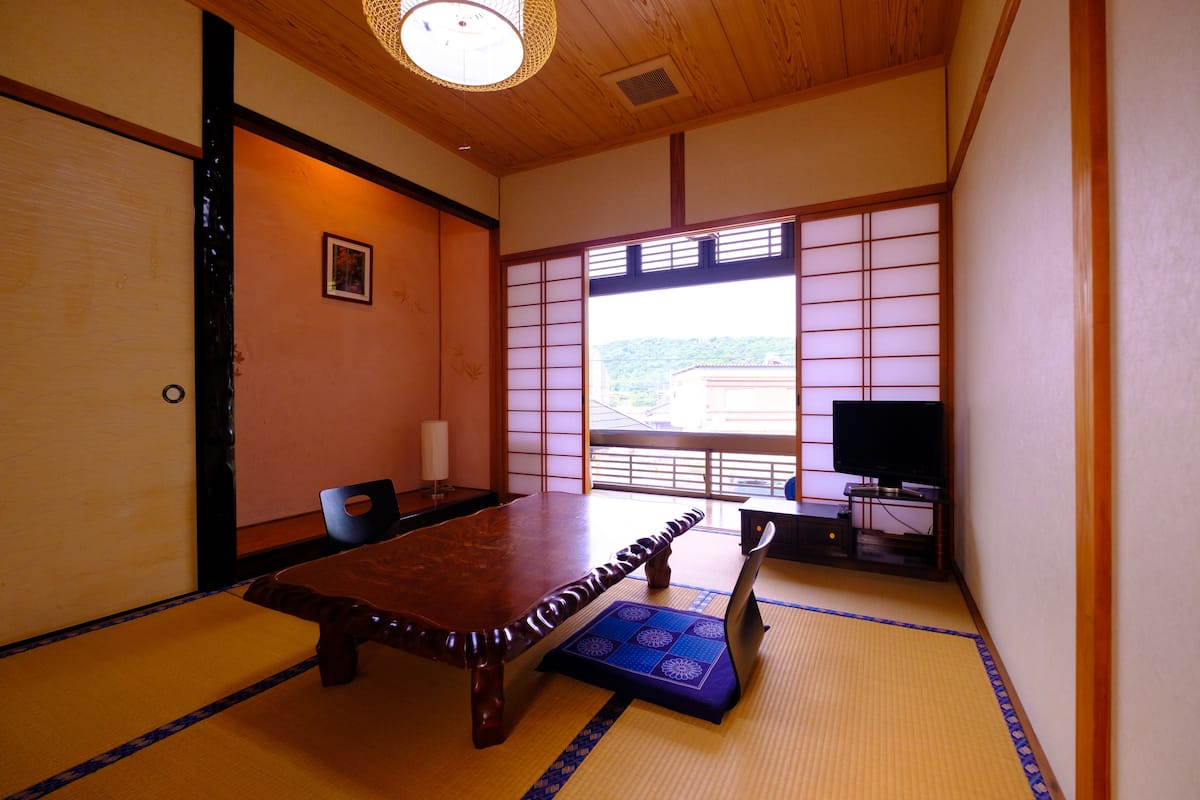 A traditional Japanese room with tatami flooring, a wooden table, a television, and a view of a hillside.
