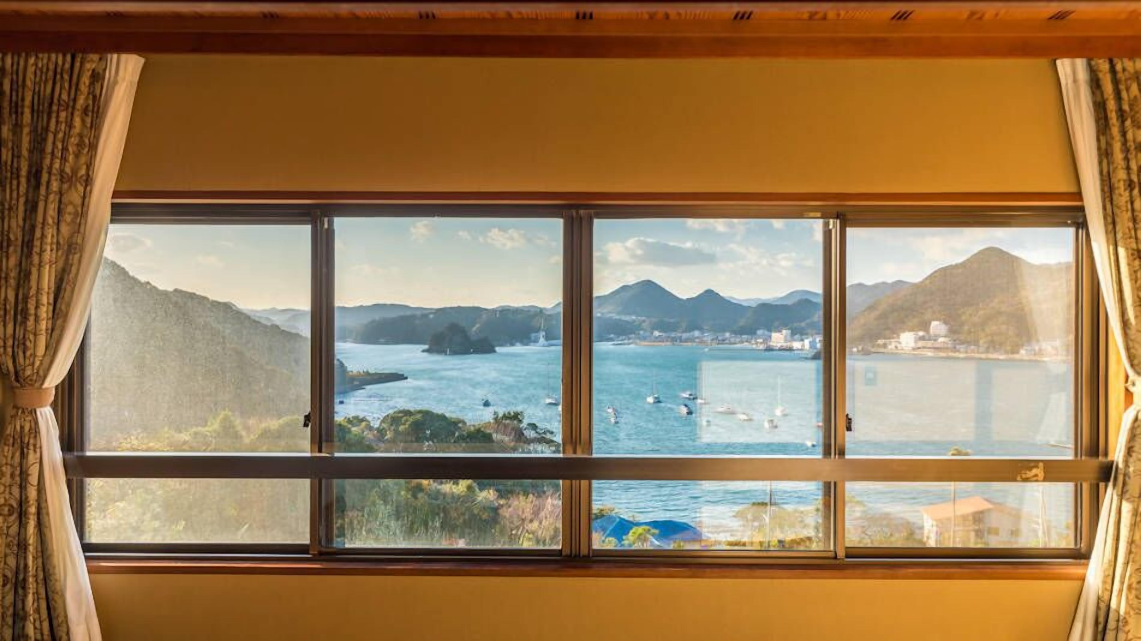 Ein Zimmer mit großem Fenster und Blick auf eine Küstenlandschaft mit Bergen und einem Gewässer.