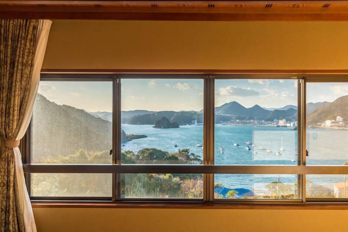 Quarto com uma grande janela que oferece vista para paisagem costeira, montanhas e um corpo de água.