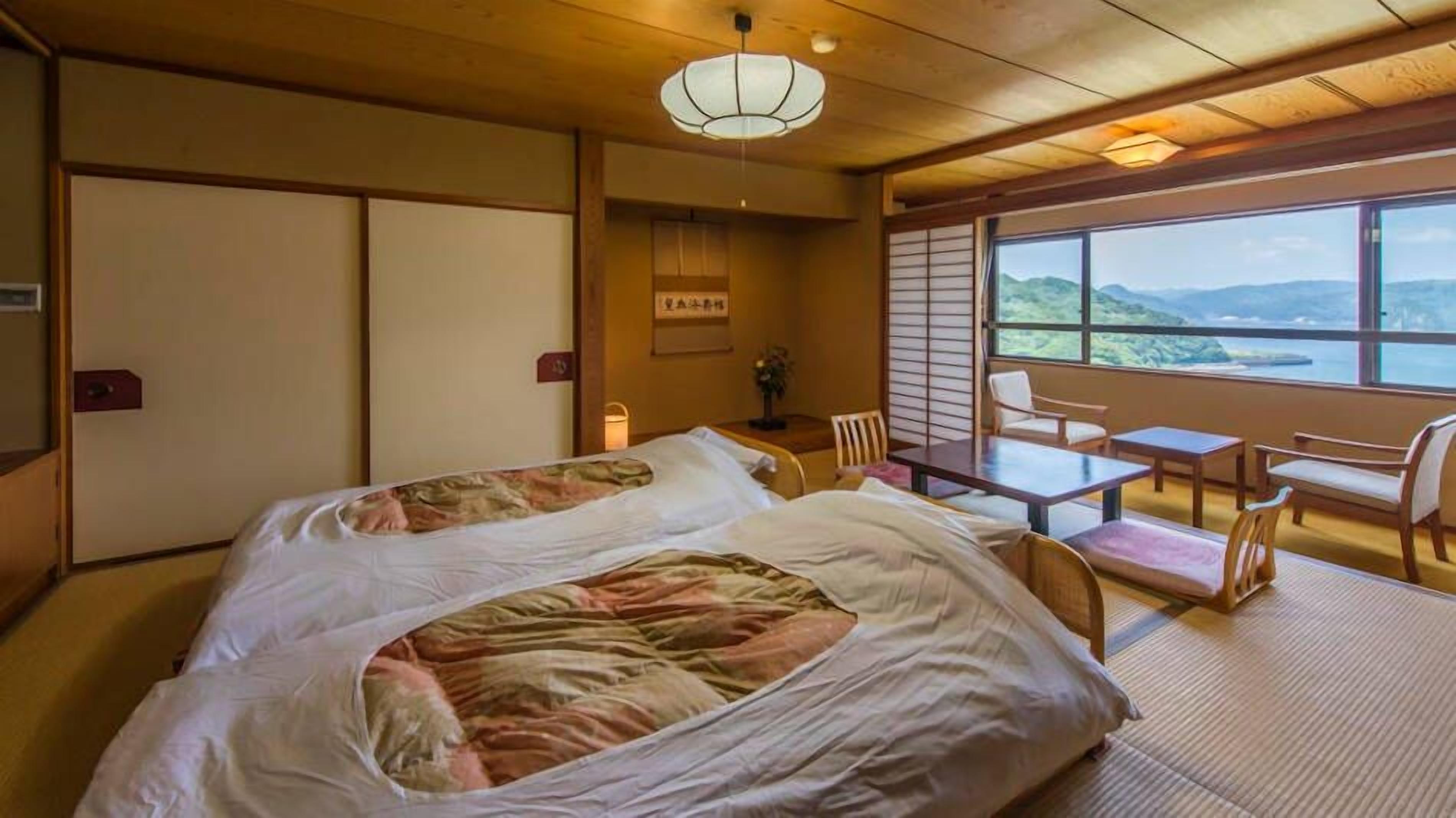 Ein traditioneller japanischer Raum mit Tatami-Böden, einem Bett mit floralem Überwurf, einem Tisch mit Stühlen und Blick auf die Berge durch große Fenster.