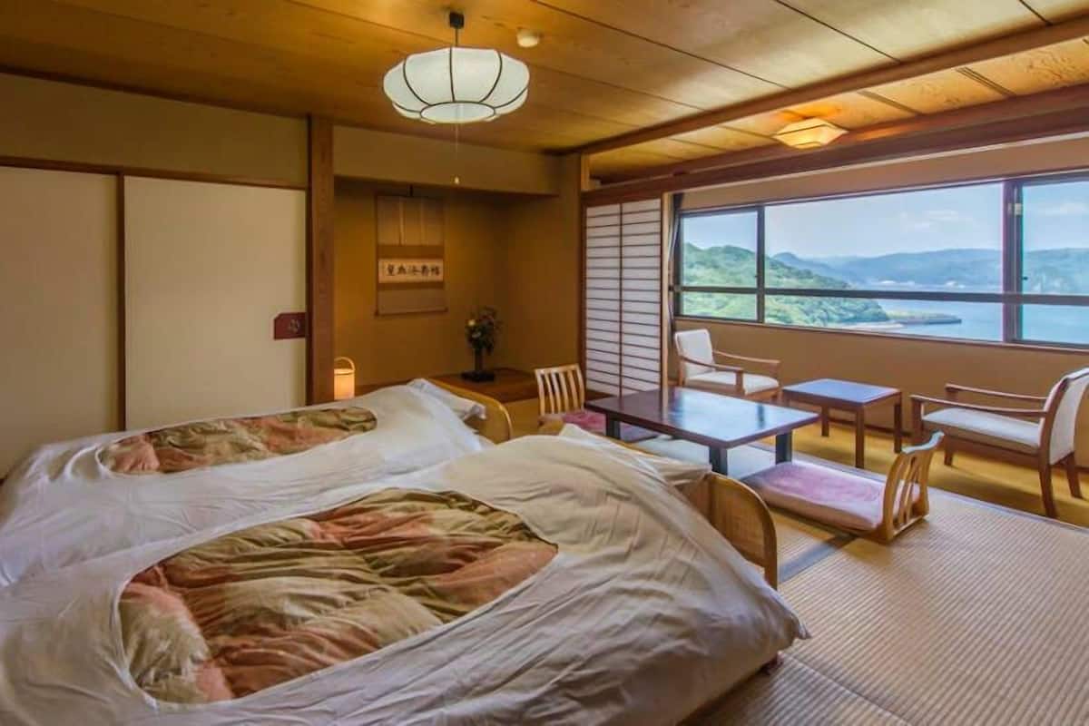 Quarto tradicional japonês com tatames, cama com colcha floral, mesa e cadeiras, e vista para as montanhas através de grandes janelas.