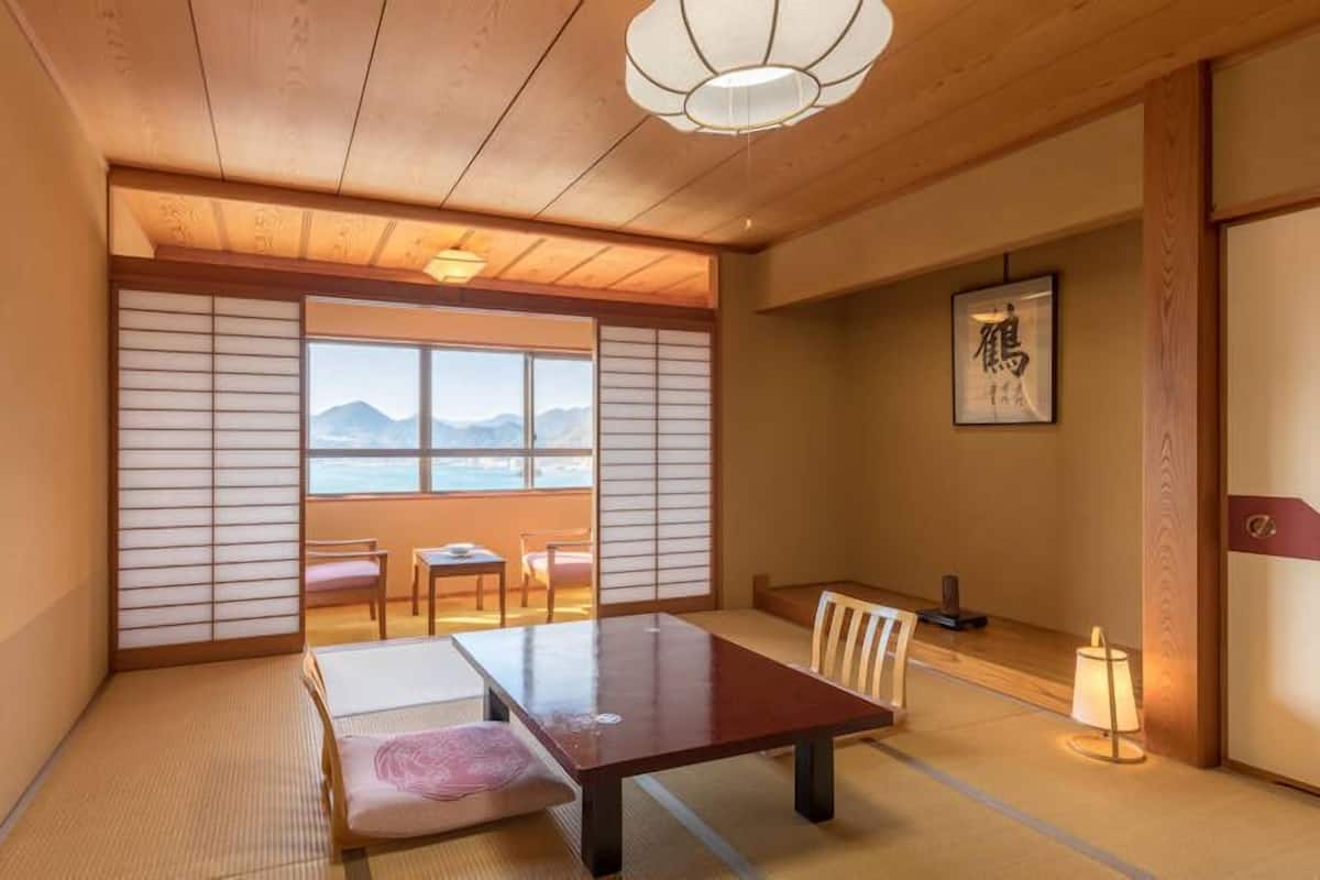 Um quarto japonês tradicional com tatames, uma mesa baixa, cadeiras, uma porta deslizante e vista para as montanhas.