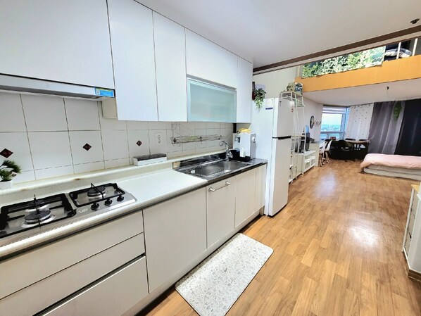 Cocina moderna con gabinetes blancos, estufa a gas y un lavamanos. Hay una pequeña zona de comedor con una mesa y sillas, y una cama al fondo.