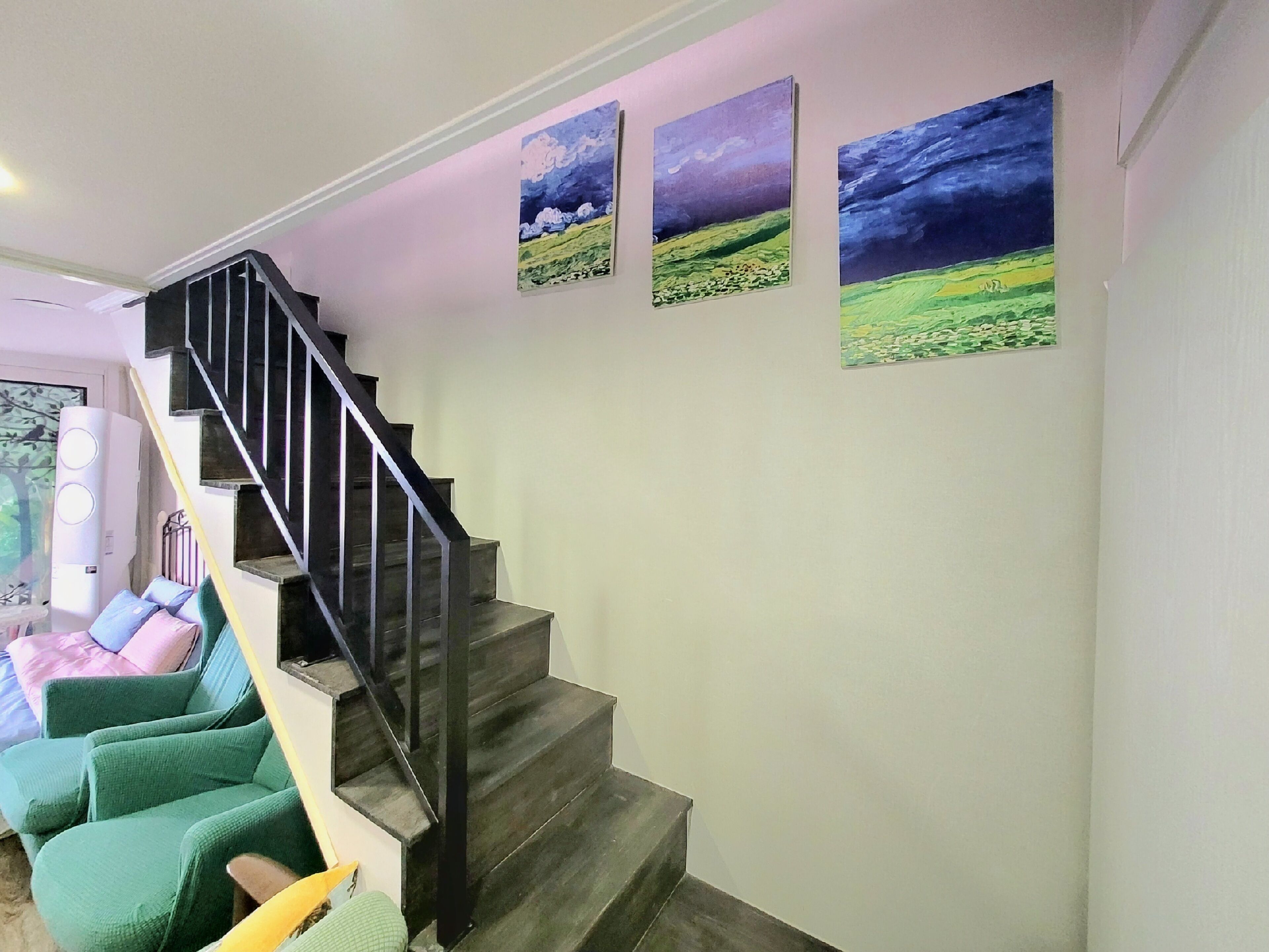 A staircase with a black railing leading to a landing. There are three paintings on the wall above the staircase depicting landscapes.
