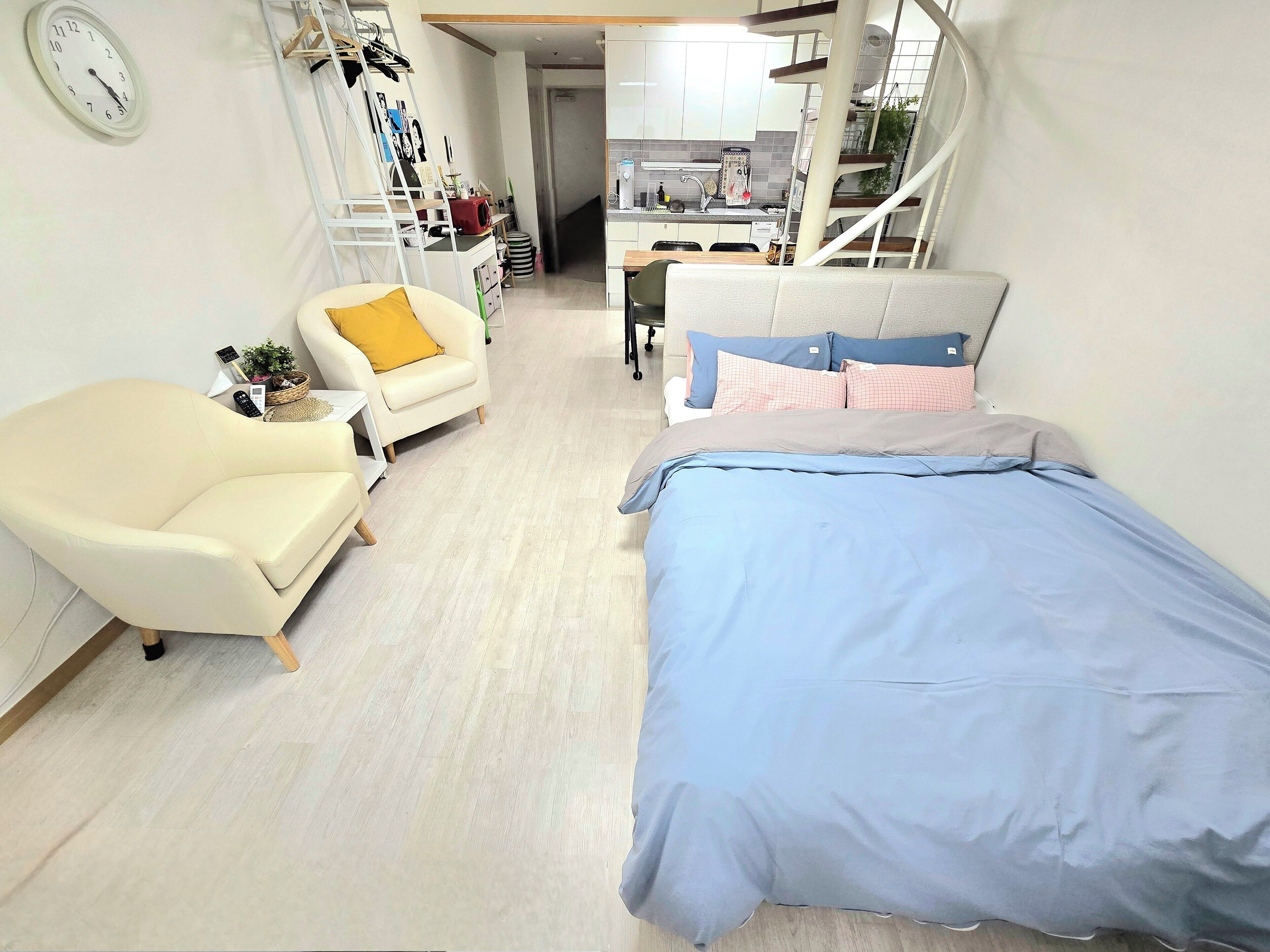 Una habitación compacta con cama, sofá y una silla. Se observa un área de cocina al fondo.