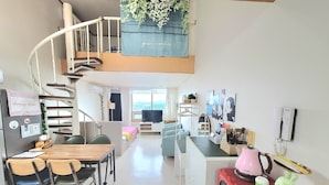 Un espacio habitable compacto con kitchenette, comedor y una pequeña zona de estar. Una escalera en espiral conduce a un nivel superior con balcón.