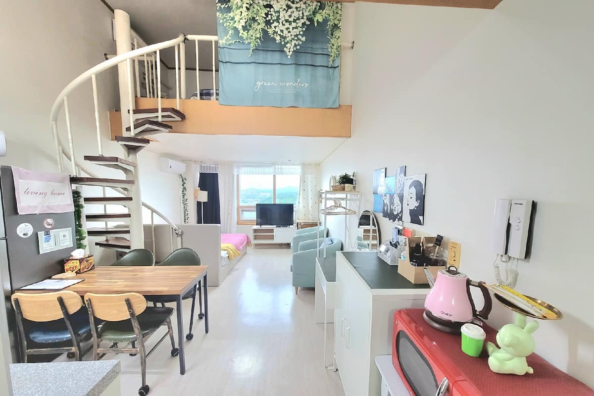 Un espace de vie compact comprenant un coin cuisine, un coin repas et un petit coin salon. Un escalier en colimaçon mène à un niveau supérieur avec un balcon.