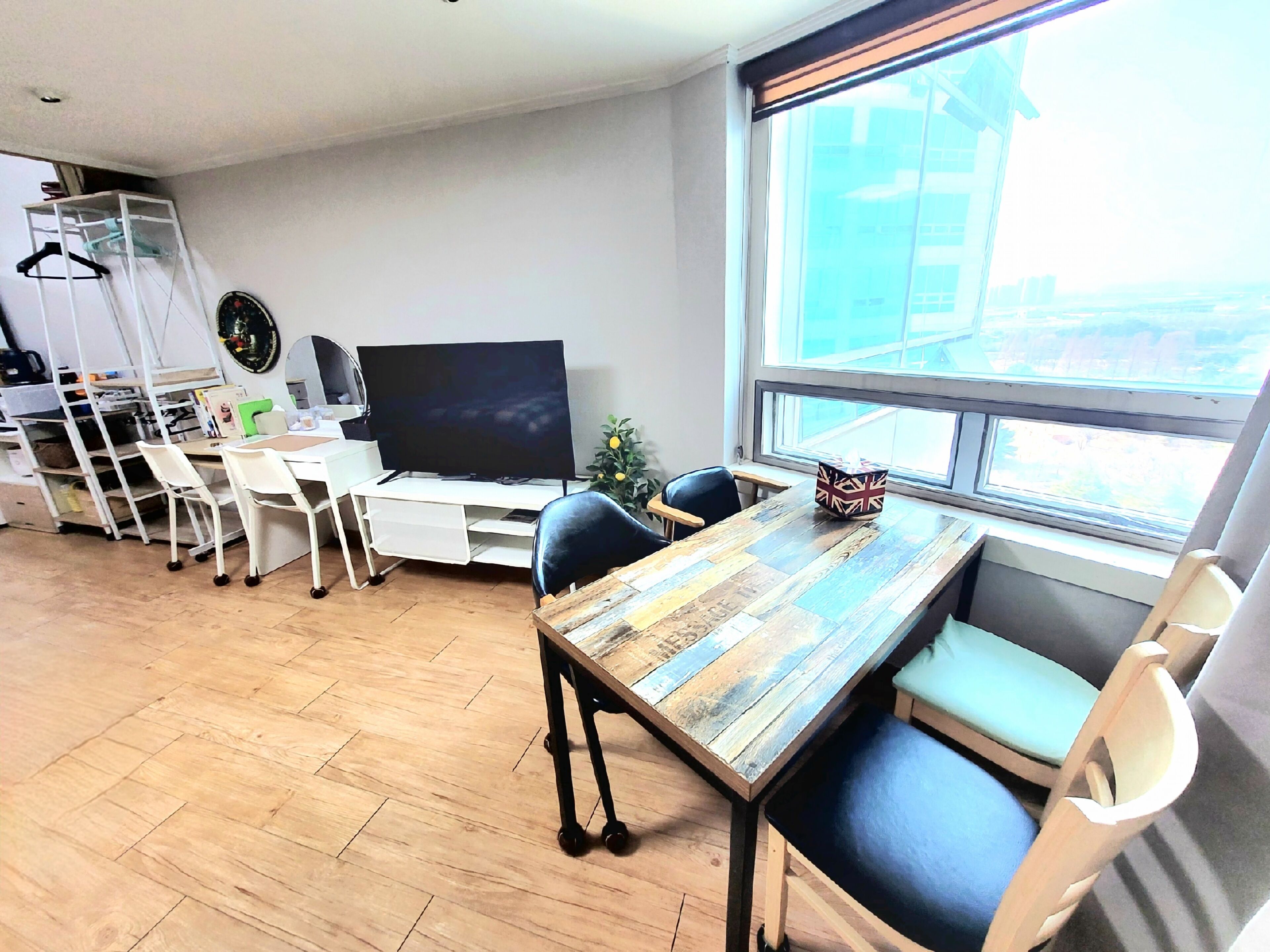 A modern living room with a wooden dining table, white chairs, a TV, and a large window with a view of buildings.