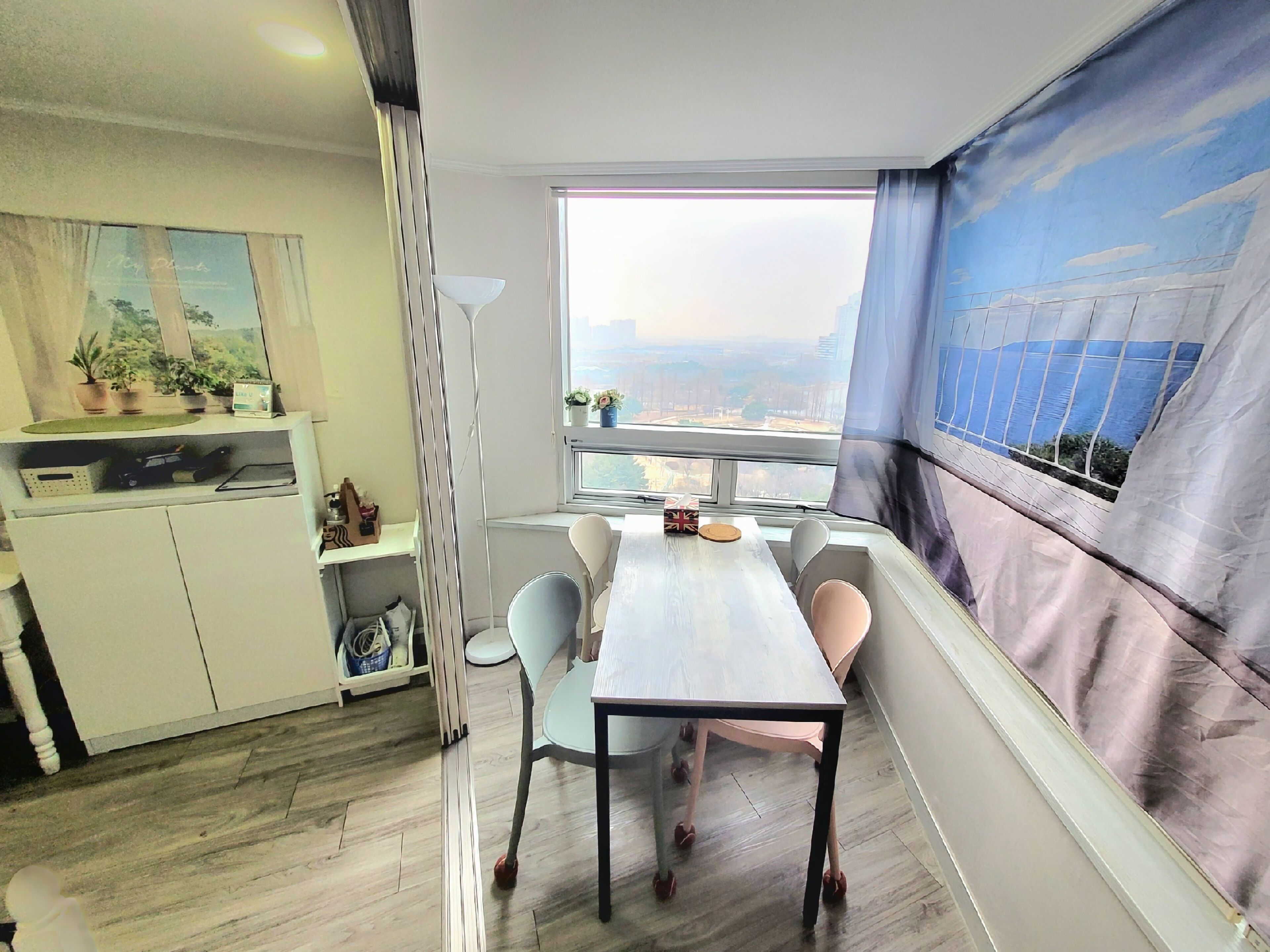 A modern room with a dining table, chairs, a window with a view of the city, a white cabinet with plants, and a curtain with a cityscape design.