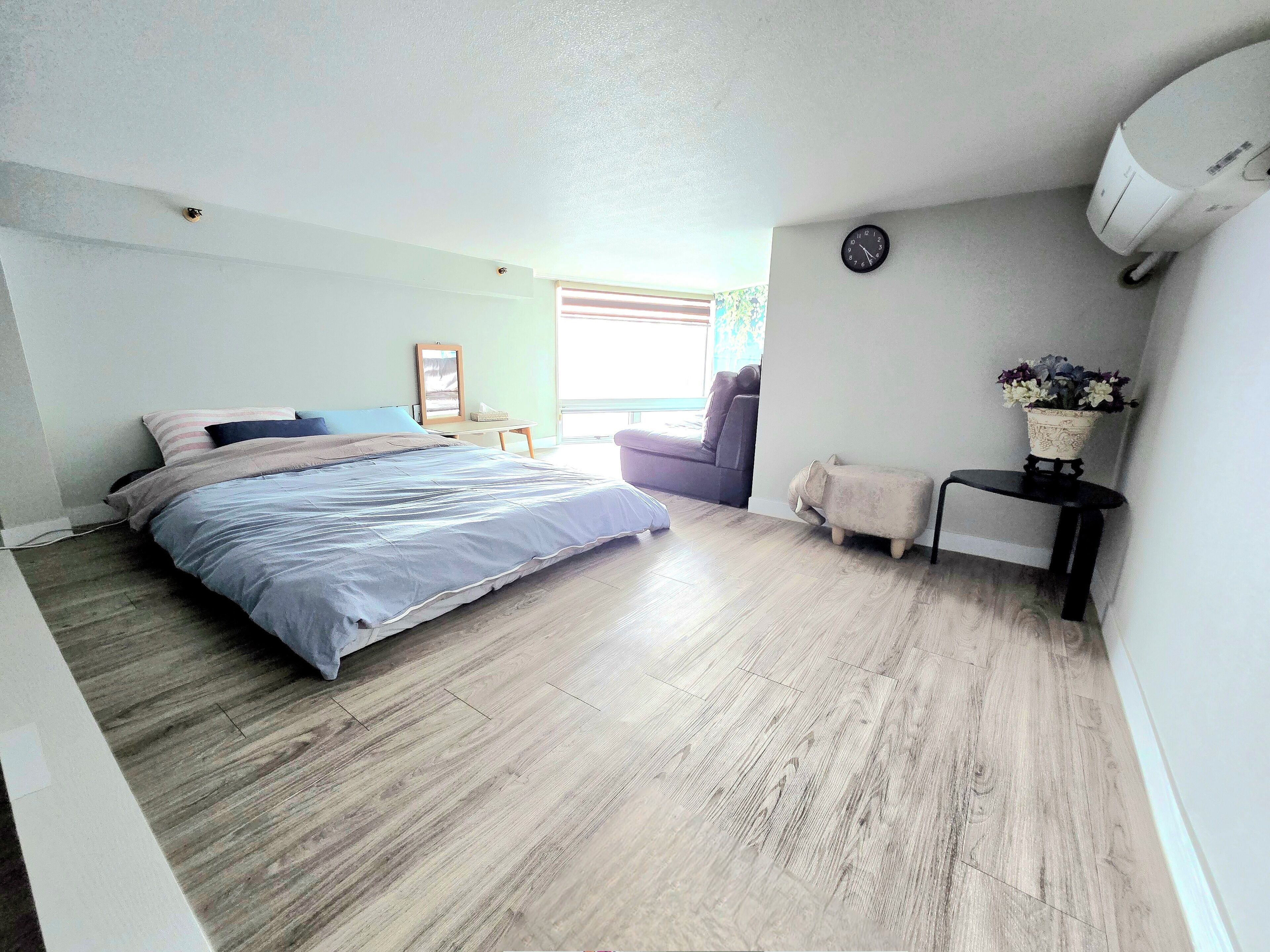 A bedroom with a bed, a small table, a chair, a clock, and a wall-mounted air conditioner.