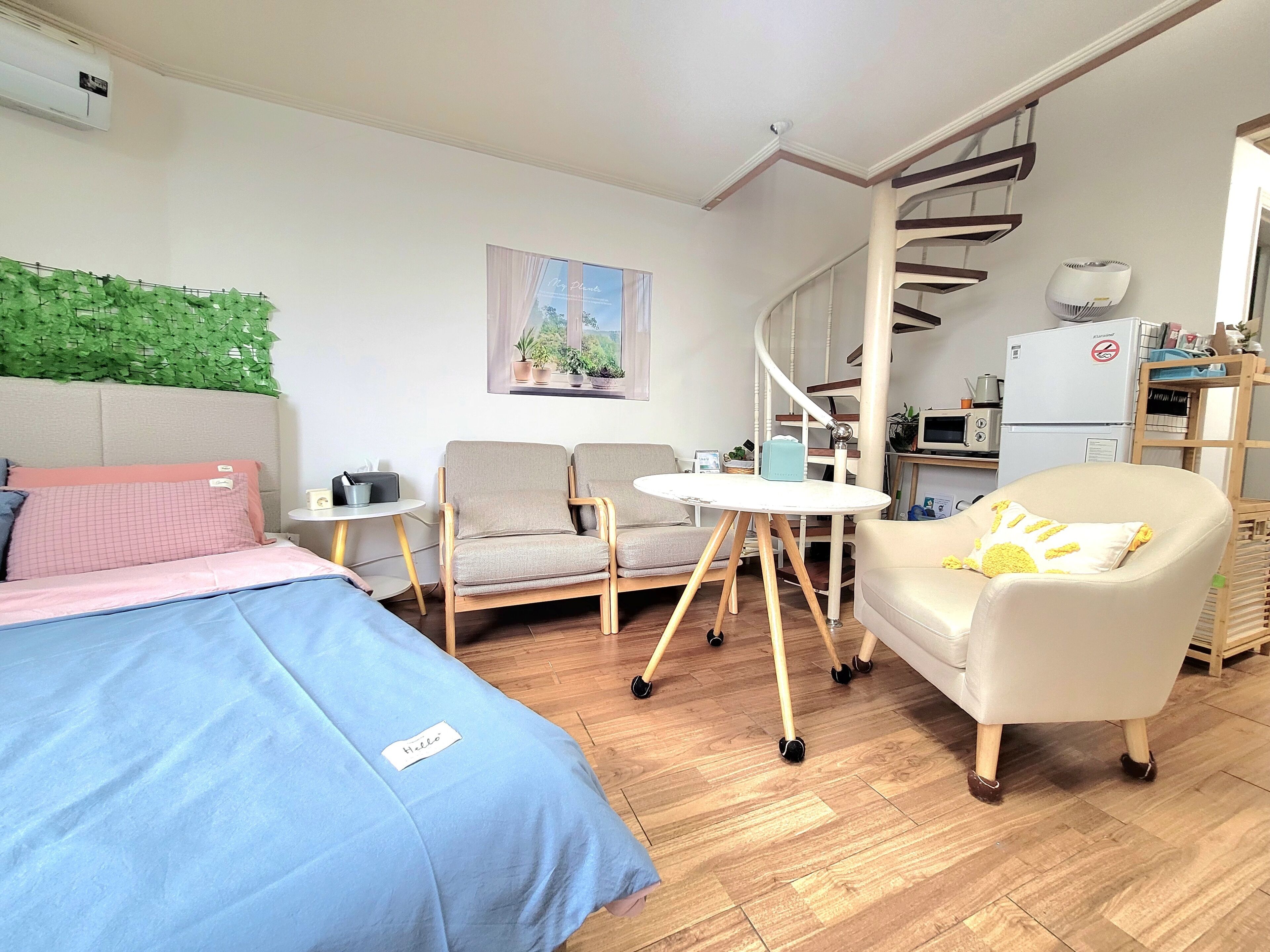 A compact room with a bed, two chairs, a small table, a sofa, a window with a view, a spiral staircase, and a kitchenette.