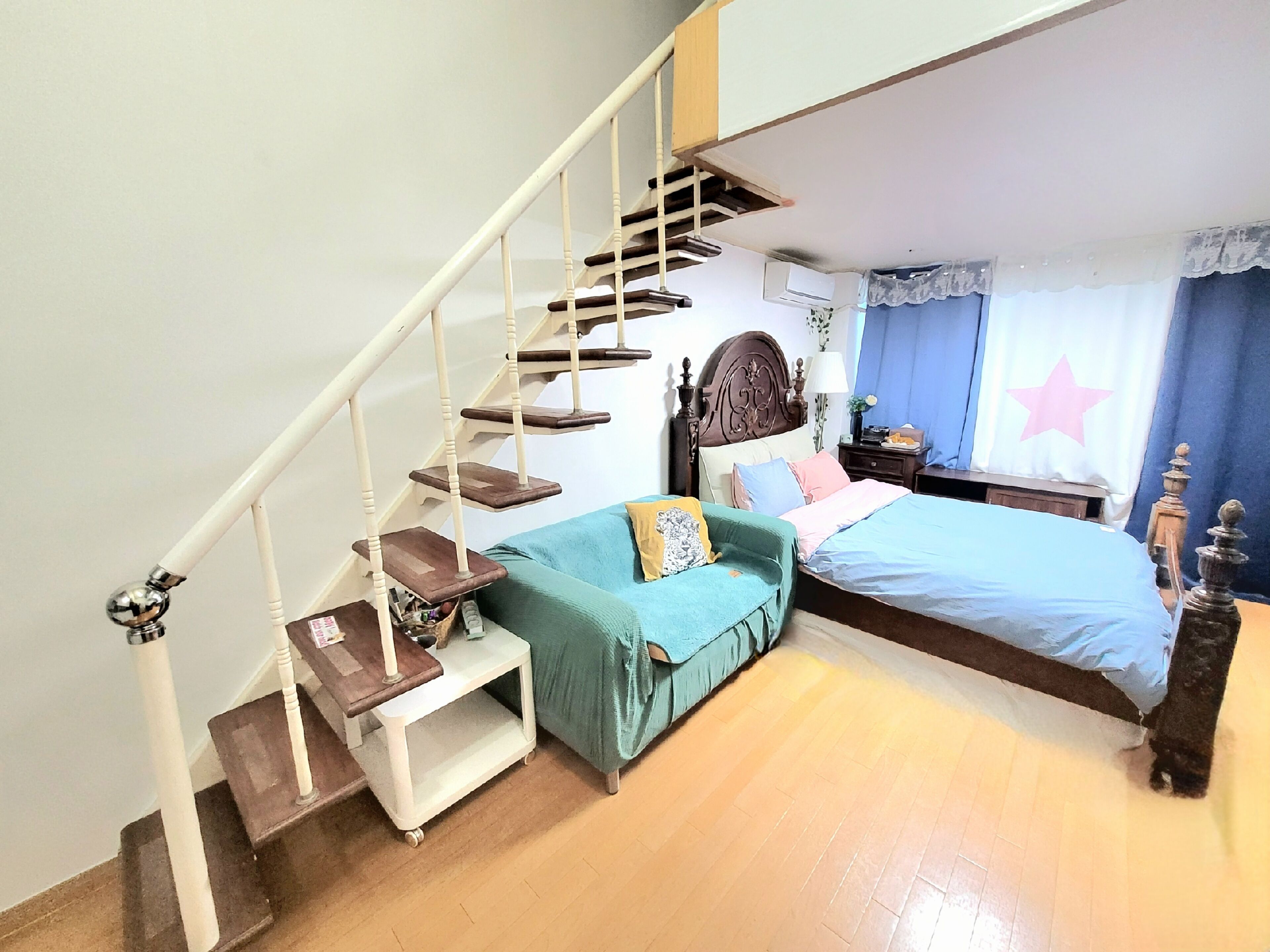 Un dormitorio con escaleras de madera, una cama con cubrecama azul, un sofá color aguamarina y una mesita de noche con una lámpara.