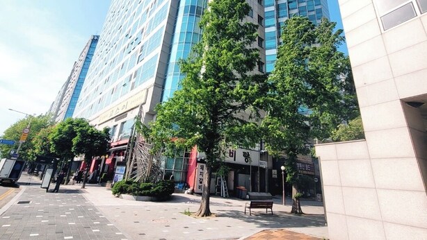 Un edificio moderno de gran altura con fachada de vidrio, rodeado por una zona peatonal con árboles y bancos.
