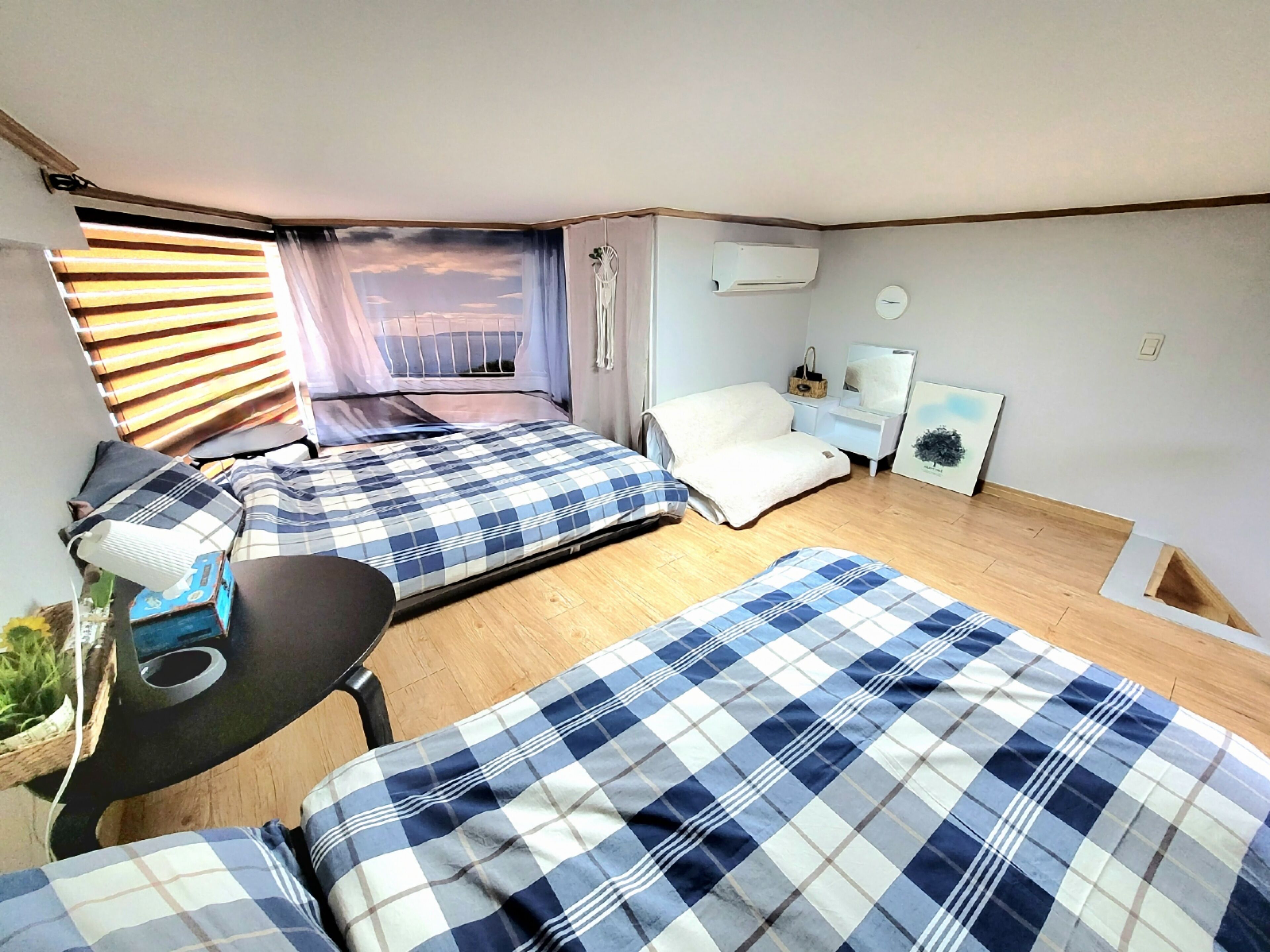 A compact room with two beds, a small table, a chair, a window with blinds, a wall-mounted air conditioner, a bedside table with a lamp, and a framed picture on the wall.