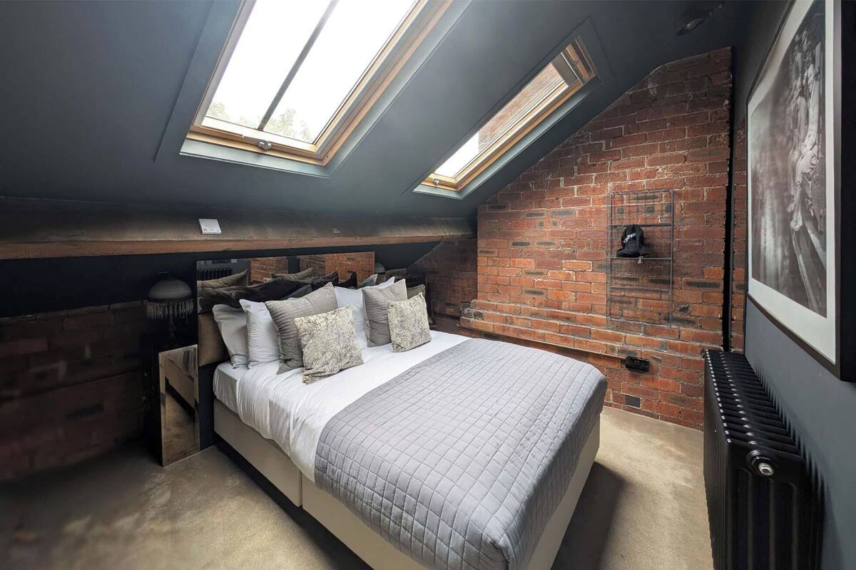 A loft bedroom with a sloped ceiling, exposed brick walls, a bed with multiple pillows, and skylights.