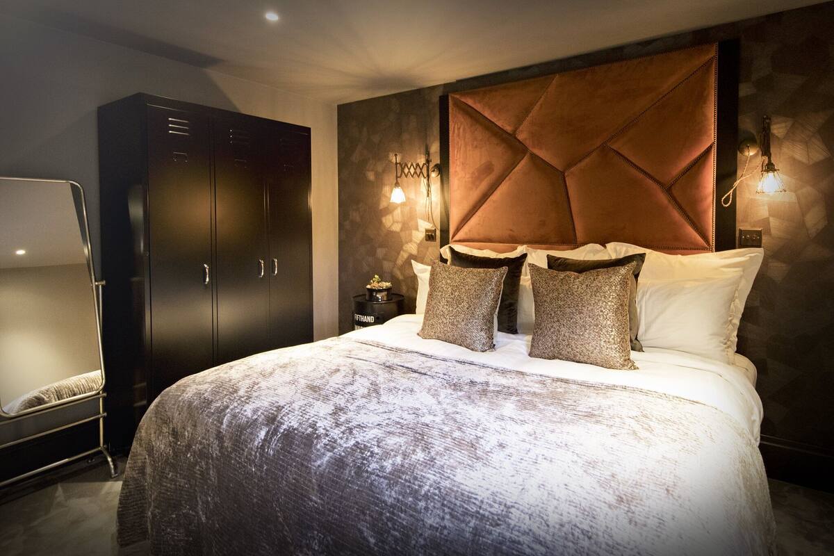 A modern bedroom with a large bed, dark wardrobe, and wall-mounted lighting.