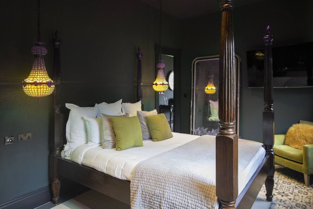 A four-poster bed with a white and beige quilt, green and white pillows, and a green chair in the background.