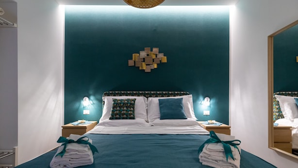 A neatly made bed with a teal bedspread and pillows, two folded towels with green ribbons, a wall-mounted light fixture, and a geometric wall decoration. - Napolilive (Naples)