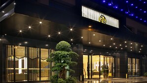 Een moderne hotelentree met een grote glazen gevel, een opvallend logo en een goed verlicht buitengebied met een boom en potplanten.