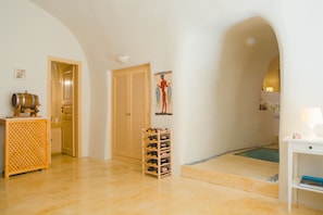 A hotel room with a wooden door, a wine rack, a small table with a lamp, and a painting on the wall. - Flora's House (Santorini)