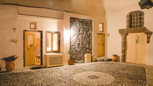 A stone-floored lobby with a central circular design, wooden doors, and a stone wall. - Flora's House (Santorini)