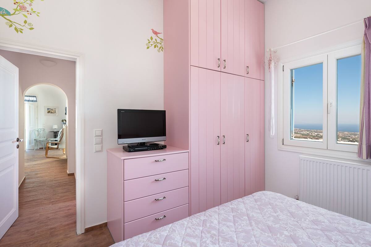 Une chambre avec une armoire rose, un lit recouvert d’une couette blanche, un téléviseur posé sur une commode et une fenêtre donnant sur un beau paysage.