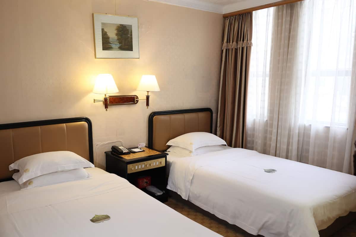 A hotel room with two beds, a nightstand with a phone and a lamp, a painting on the wall, and a window with curtains.