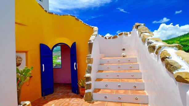 Una escalera que lleva a una puerta con marco azul, rodeada de paredes blancas y un exterior amarillo.