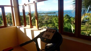 A room with a stationary bike, large windows with a view of a lush landscape and a body of water. - Oasis Lodge (Fenoarivo Atsinanana)