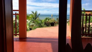 A view from a covered outdoor area with a concrete floor, leading to a pathway surrounded by tropical plants and a view of the ocean. - Oasis Lodge (Fenoarivo Atsinanana)