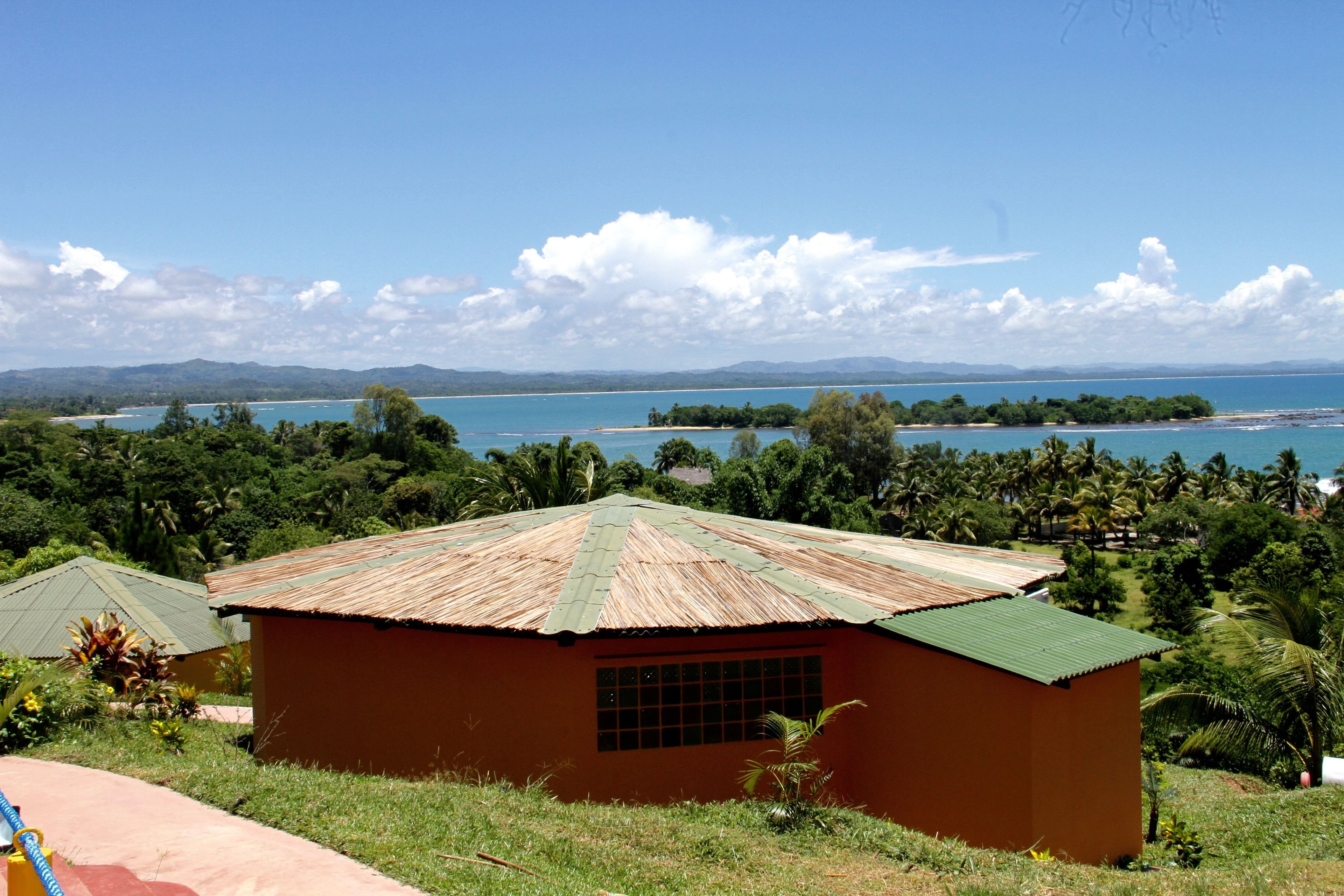 A small, single-story building with a green roof, surrounded by lush greenery and a view of a large body of water and distant mountains.