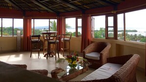A room with a high ceiling, large windows, a dining table with chairs, and wicker furniture. - Oasis Lodge (Fenoarivo Atsinanana)